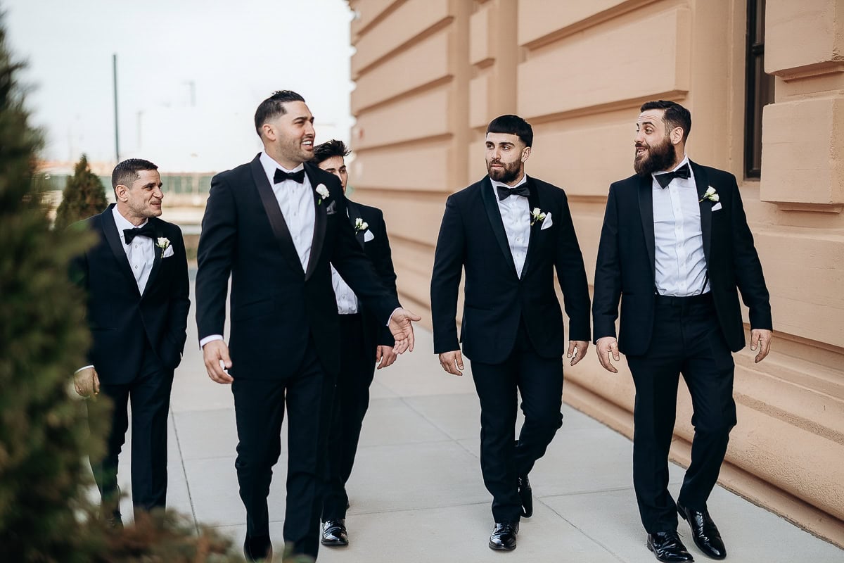 Five men in black tuxedos with boutonnières walk past a limestone facade outside The Switch House by Cescaphe in Philadelphia.