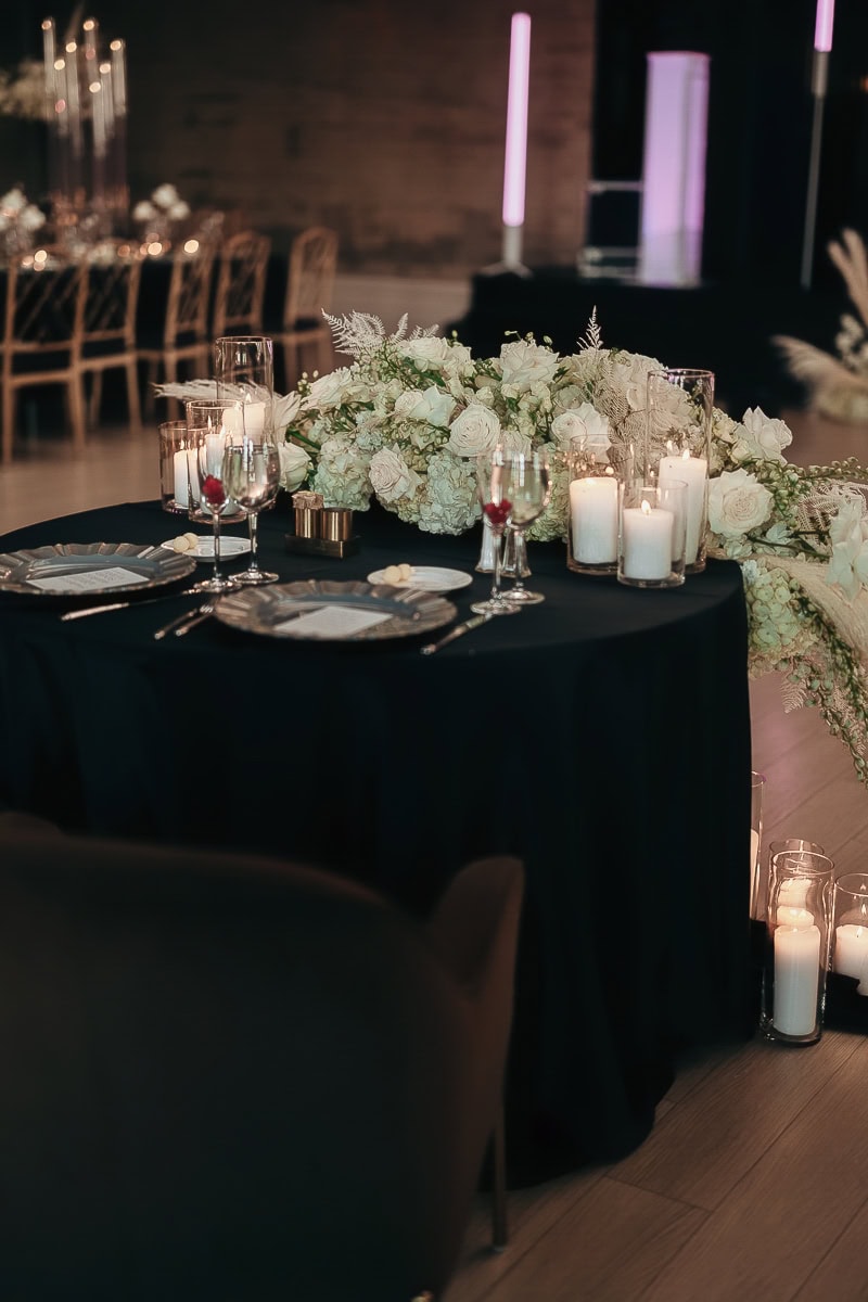 Sweetheart table with black linen, ivory rose–hydrangea garland, glass pillar candles, and champagne flutes; purple uplighting at The Switch House.