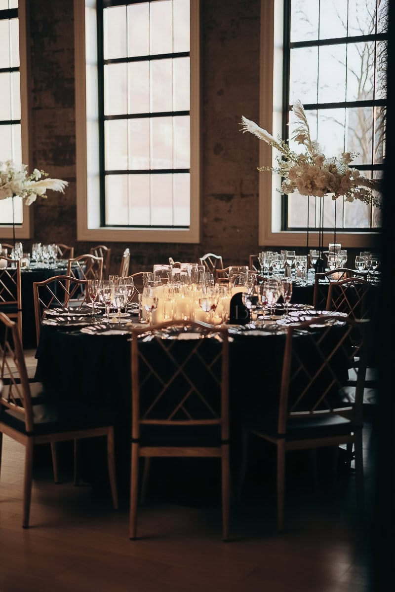 Candlelit round table with black linens, crystal stemware, and tall white florals by industrial windows at The Switch House, Philadelphia.