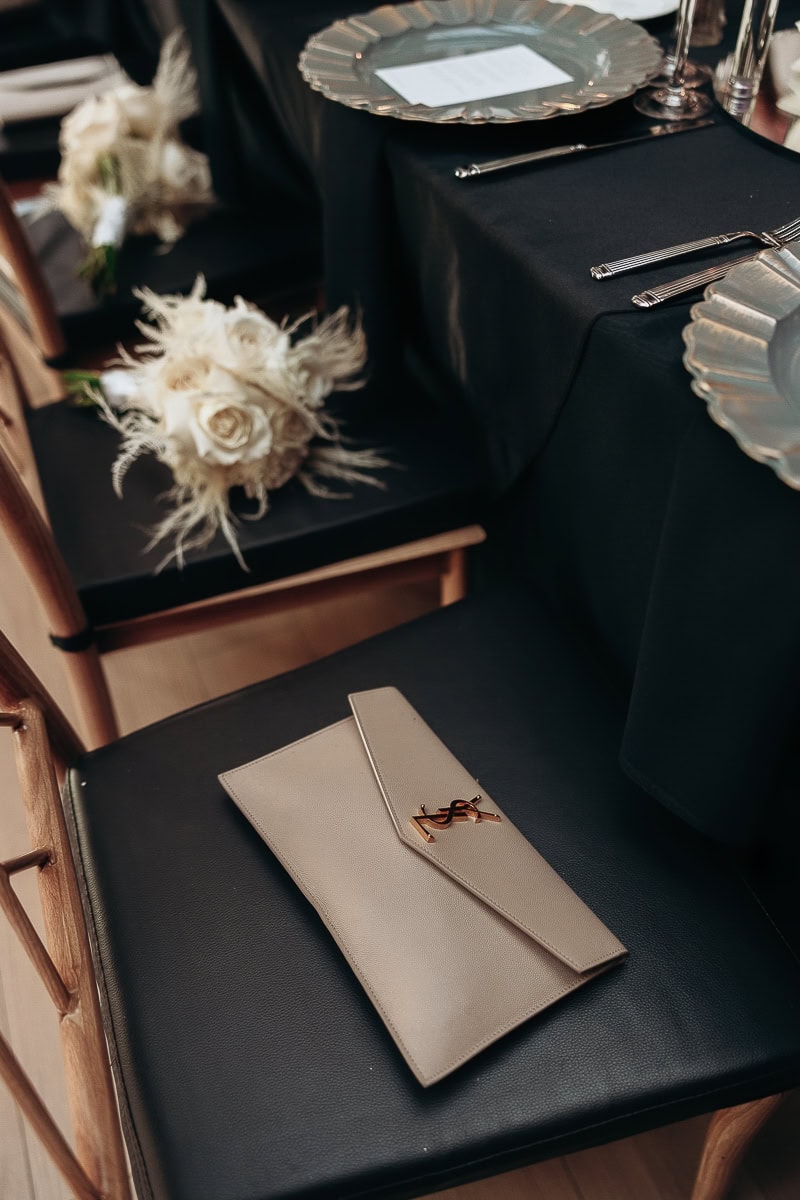 Ivory YSL clutch on black leather chair beside white rose bouquet and black-linen place setting with scalloped charger at The Switch House, Philadelphia.