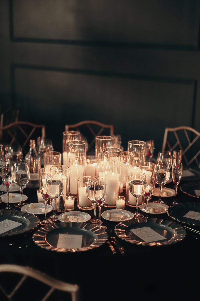 Pillar candles in glass hurricanes glow over a black-linen wedding table with ruffled chargers, bread plates, and crystal stemware at The Switch House by Cescaphe.