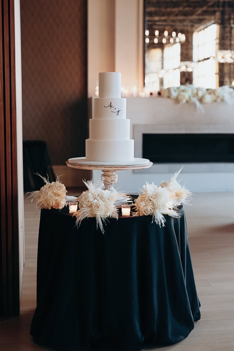 Four-tier white wedding cake with A+J monogram on vintage stand, surrounded by ivory rose bouquets and votives on black velvet table.