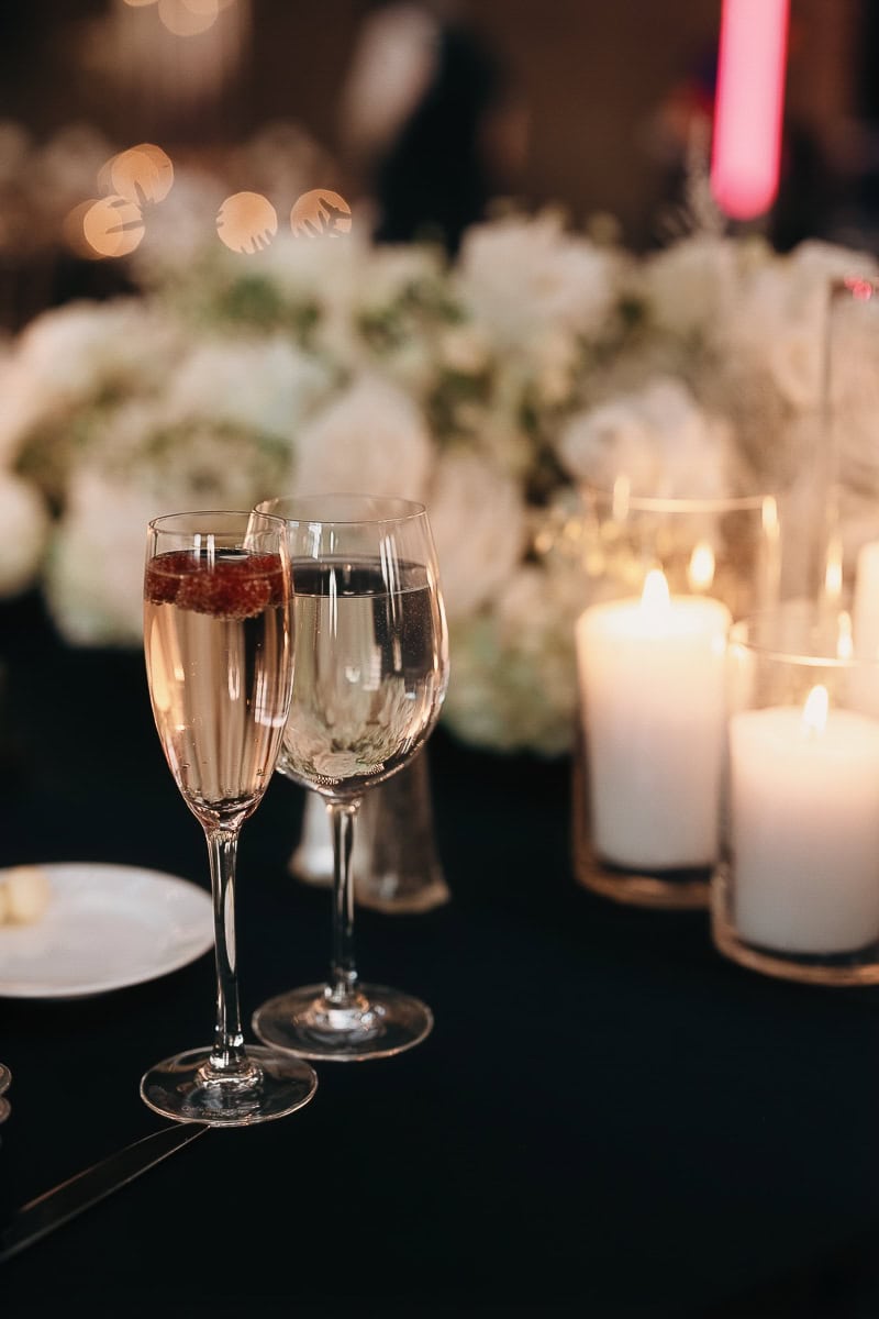 Wedding reception table: champagne with raspberry, water glass, white pillar candles, and white‑rose centerpiece on black linen.
