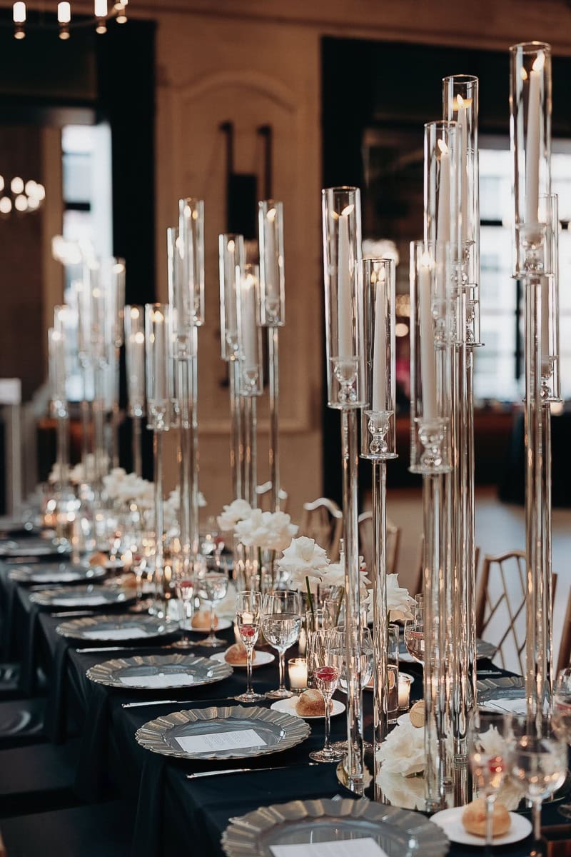 Long black-linen banquet table with tall glass tapers, white florals, silver chargers, and crystal stemware at The Switch House by Cescaphe.