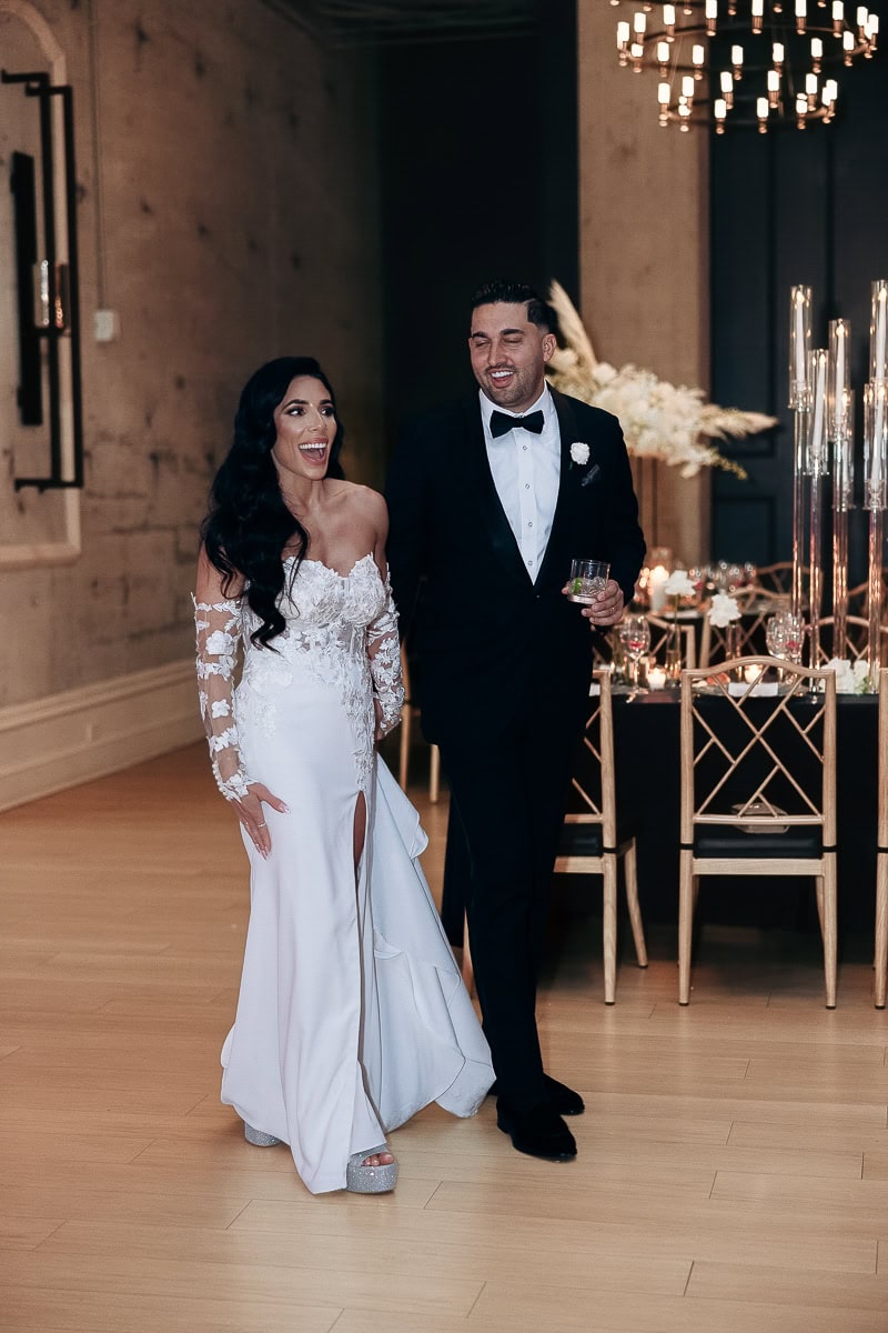 Bride in lace-sleeved gown and groom in black tux walk into a candlelit reception with tall candles and white florals at The Switch House.