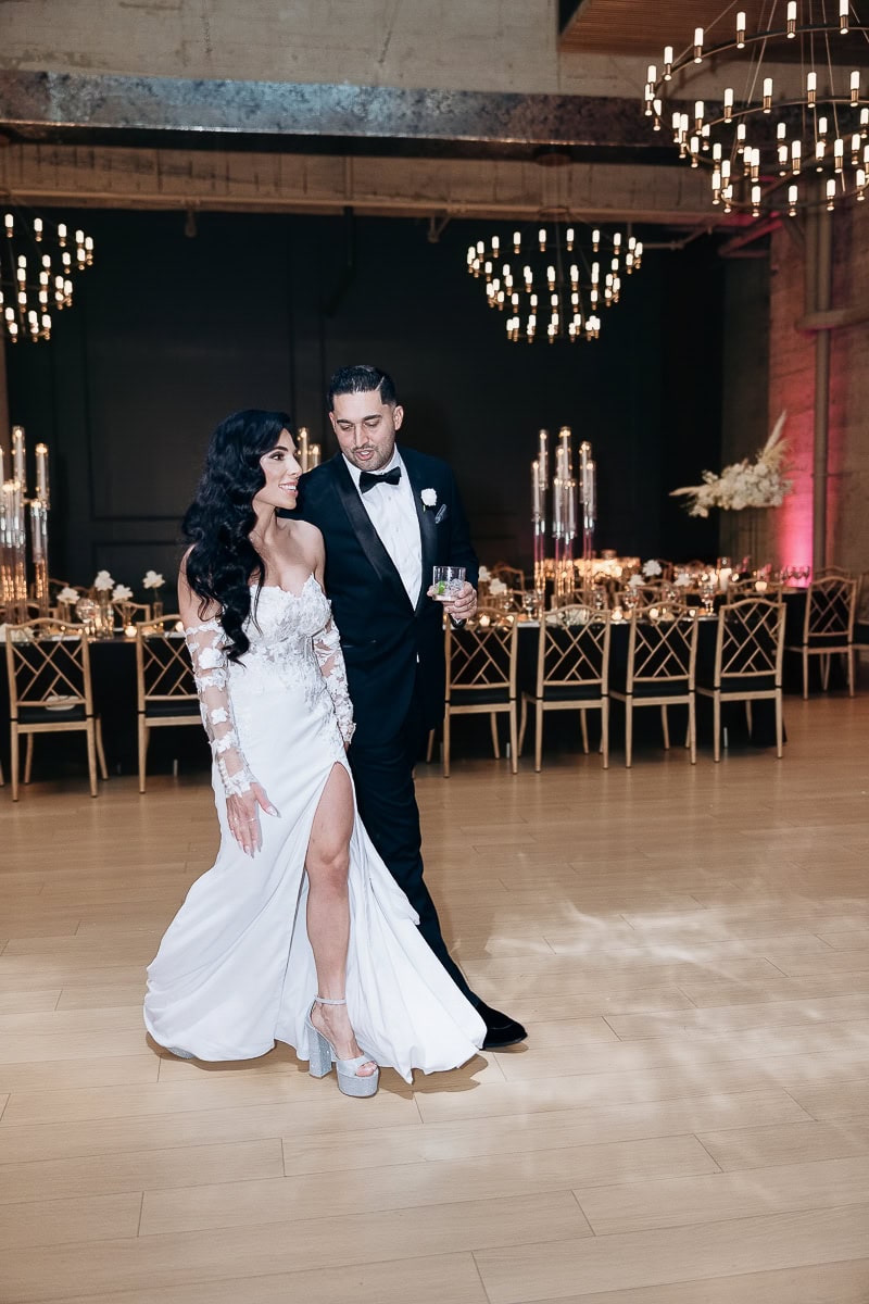 Couple in lace-sleeve gown and tux walk the dance floor under chandeliers beside candlelit tables at The Switch House, Philadelphia.