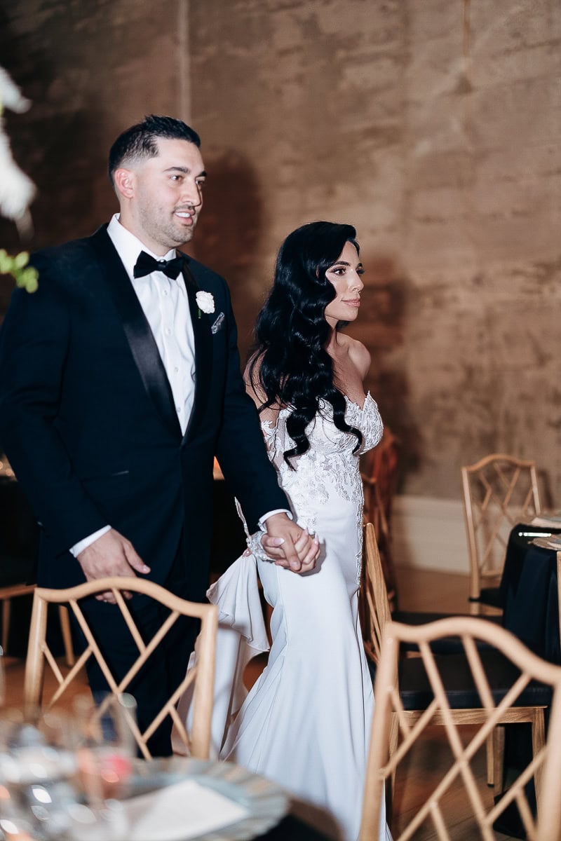 Bride in off-shoulder lace crepe gown and groom in black tux walk hand-in-hand past black linens and wood chairs at The Switch House, Philadelphia.