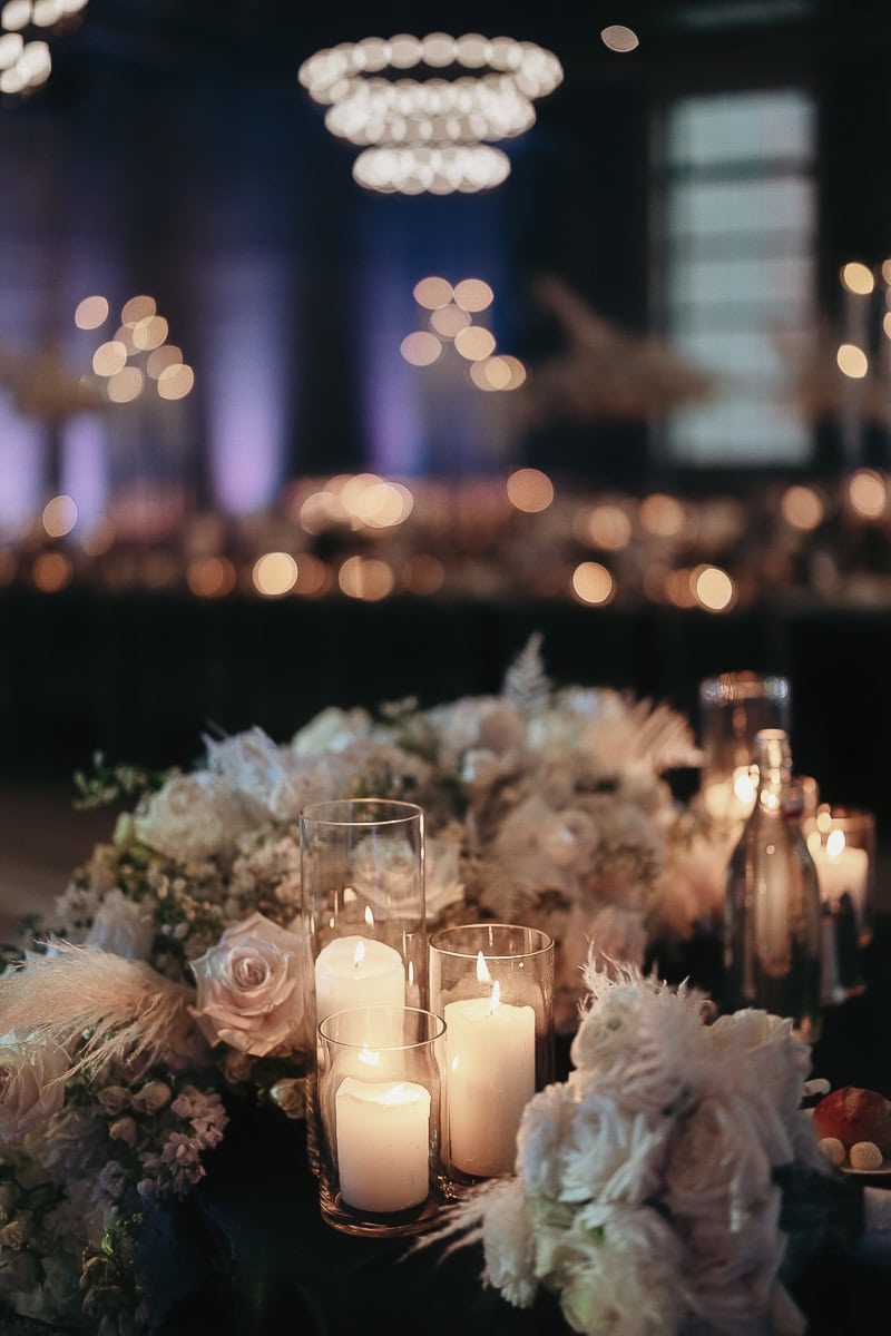 Pillar candles in glass hurricanes beside ivory florals on a reception table; purple uplighting and ring chandeliers softly blurred in the background.