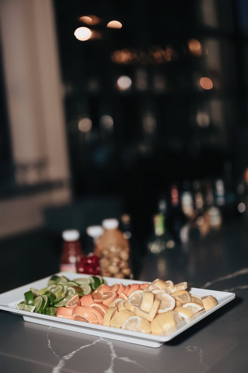 White tray of lemon, lime, and orange garnishes on a dark marble bar with blurred bottles and lights in the background.