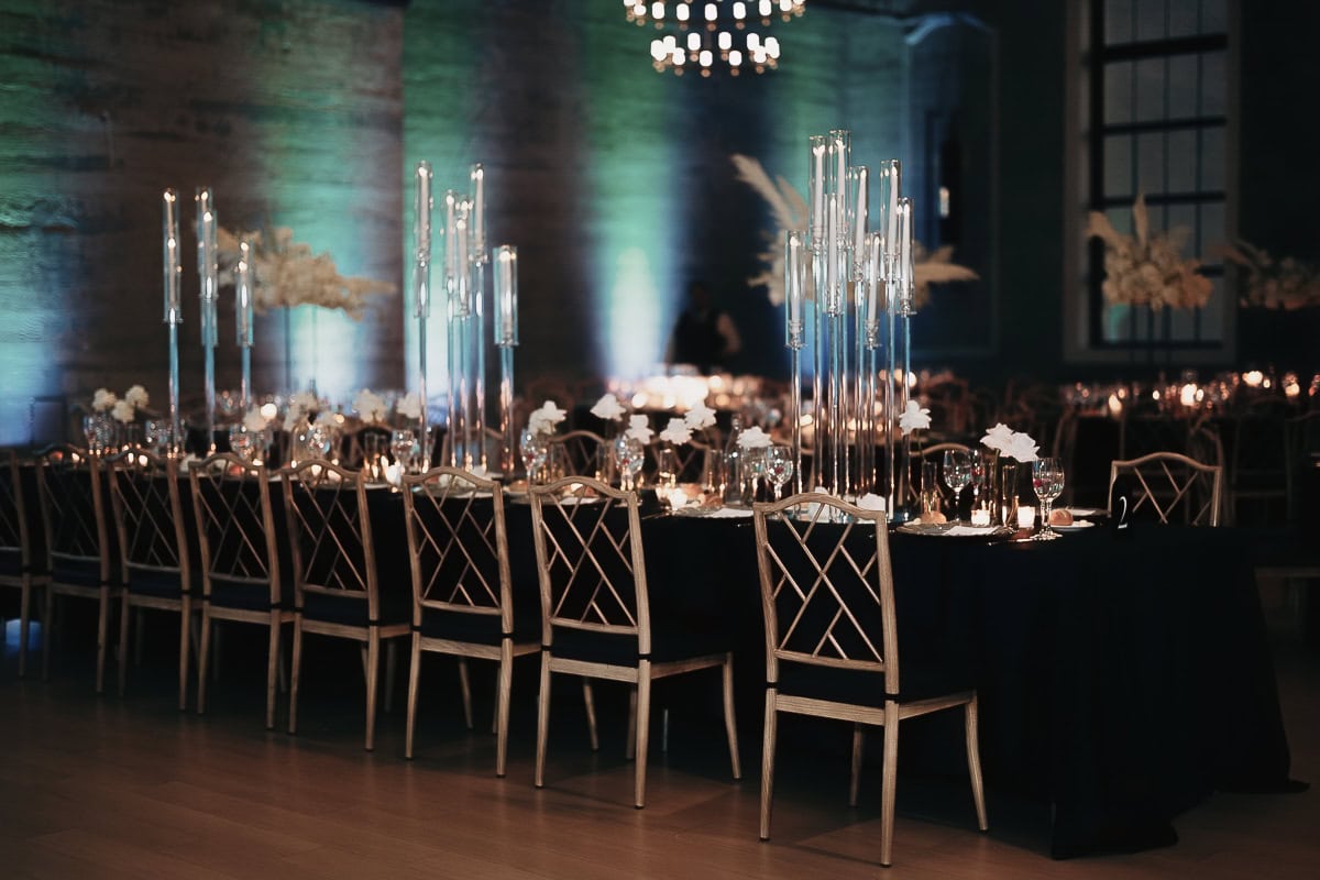 Long black-clad banquet tables with tall glass candle cylinders, white bud vases, and blue uplighting at The Switch House by Cescaphe.