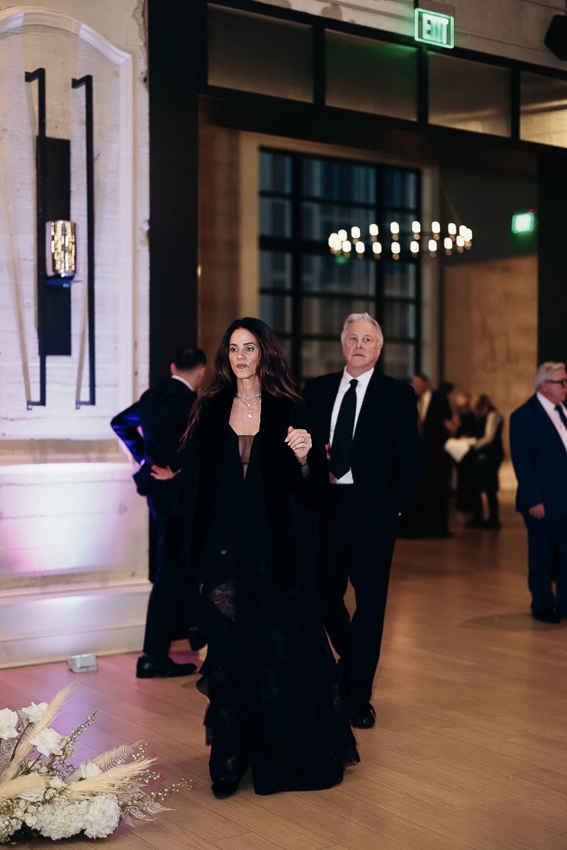 Guests in black-tie walk through lilac-lit reception space with modern sconces, chandelier bokeh, and a white rose–pampas floral arrangement on the floor.