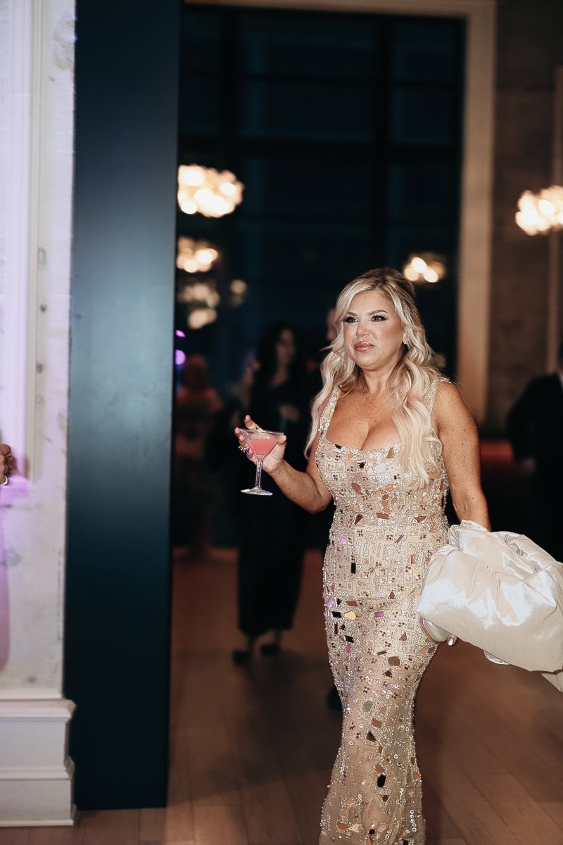Guest in mirrored sequin gown walks with pink cocktail and satin coat; soft chandeliers behind at The Switch House, Philadelphia.