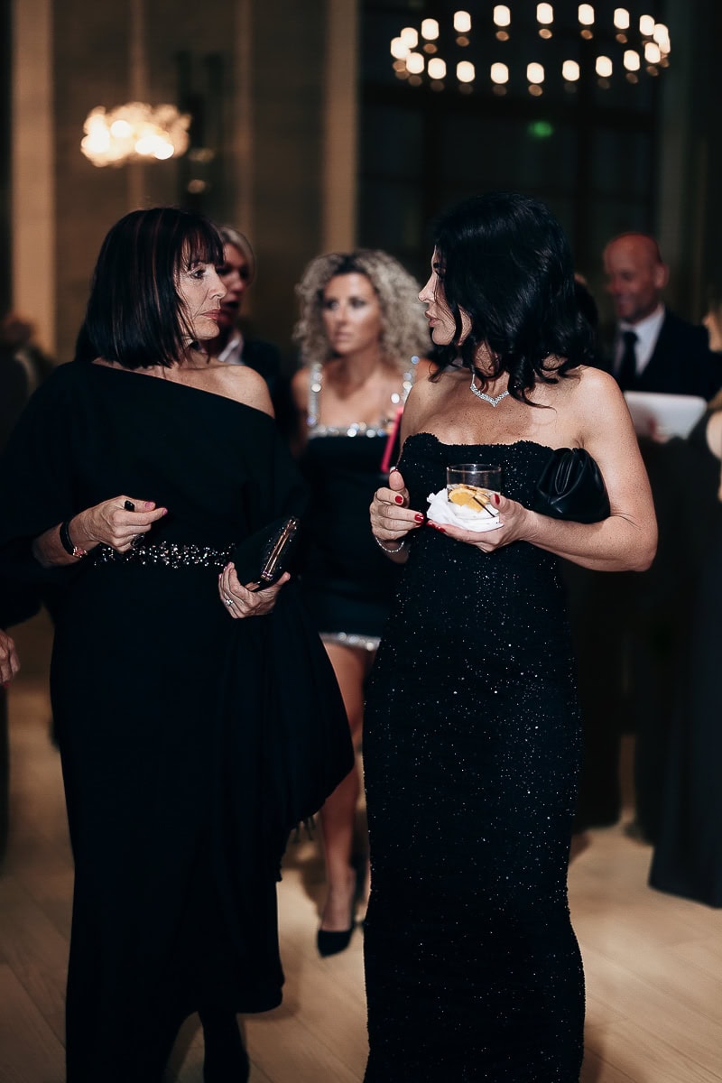 Two guests in black gowns talk at a wedding reception; one holds a lemon cocktail; blurred chandeliers in background at The Switch House.