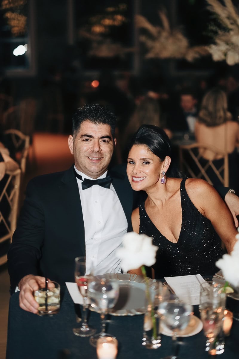 Two guests in a tuxedo and black sequin dress sit at a candlelit reception table with cocktails and white roses at The Switch House, Philadelphia.