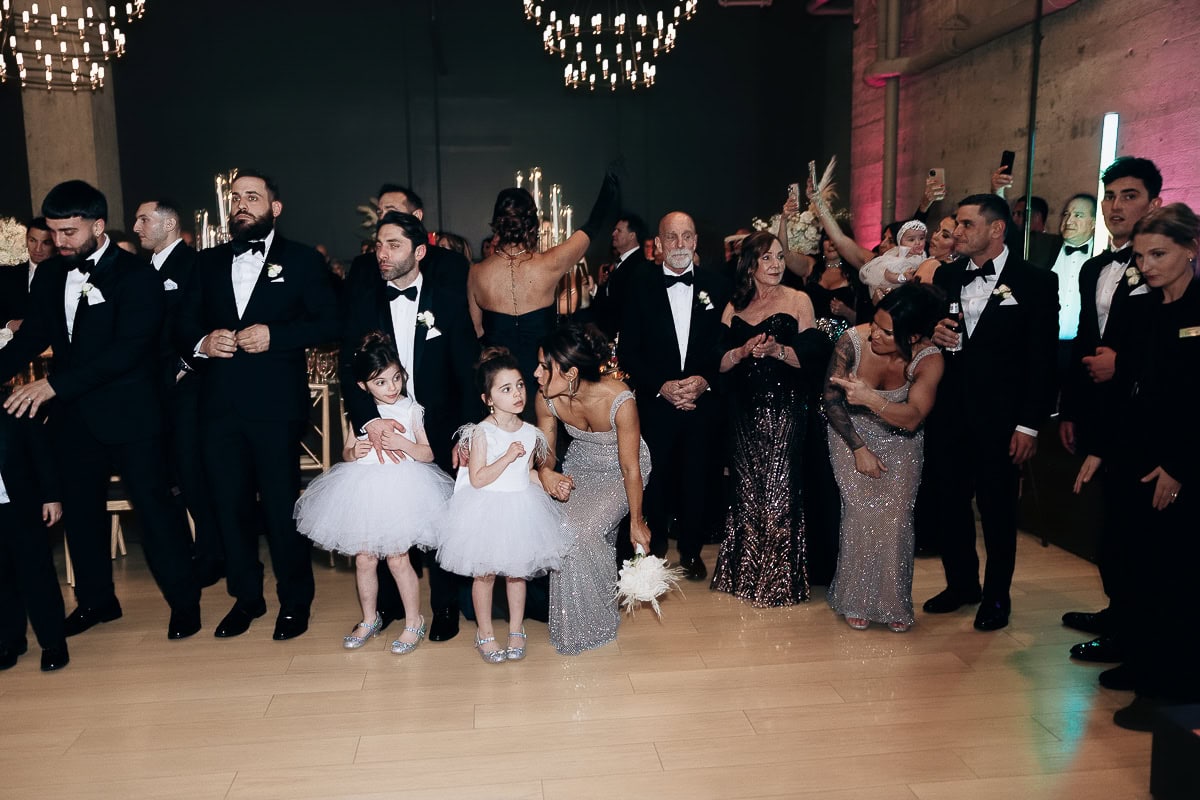 Black‑tie guests with two flower girls pause on the dance floor under chandeliers at The Switch House by Cescaphe, Philadelphia.