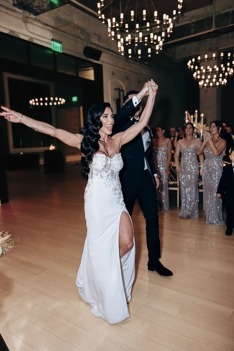 Couple enter reception; bride in strapless slit gown, chandeliers above, guests in sequined dresses applaud at The Switch House, Philadelphia.