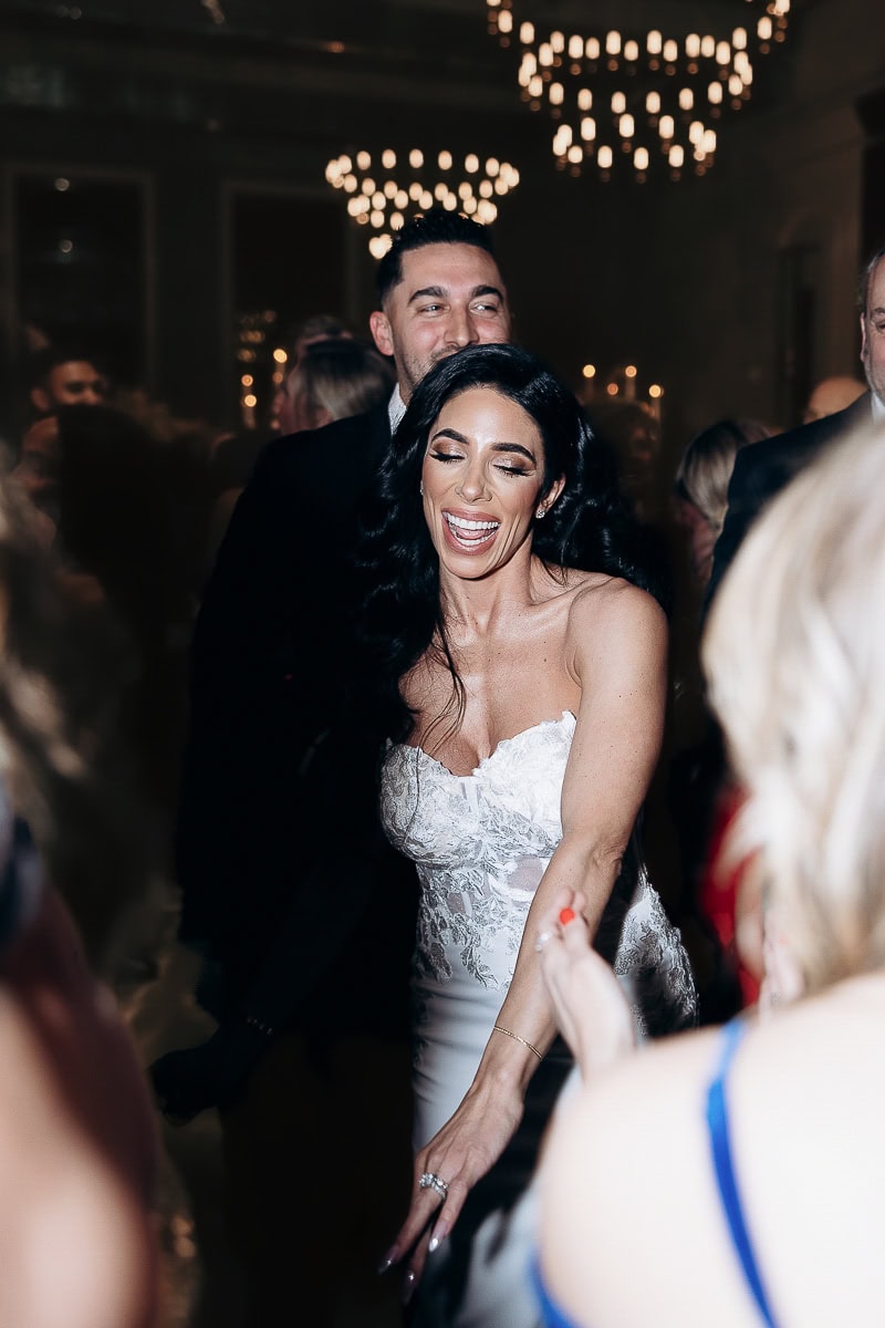 Bride in lace strapless gown dances under chandeliers; partner behind amid crowd, flash-lit. The Switch House by Cescaphe, Philadelphia.