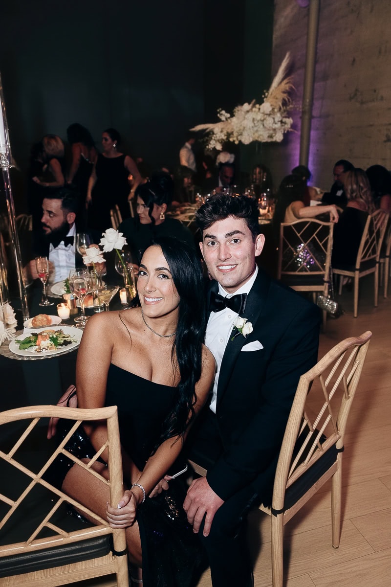 Two wedding guests at a candlelit reception; one in a black gown, one in a tux with white boutonniere; glassware and floral centerpiece visible.