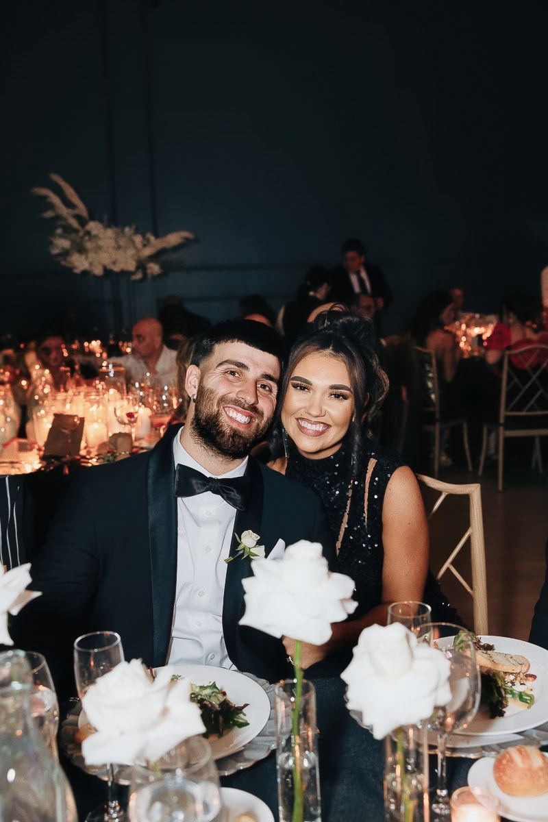 Two black-tie guests smiling at a candlelit reception table with white roses and glassware at The Switch House by Cescaphe, Philadelphia, PA.