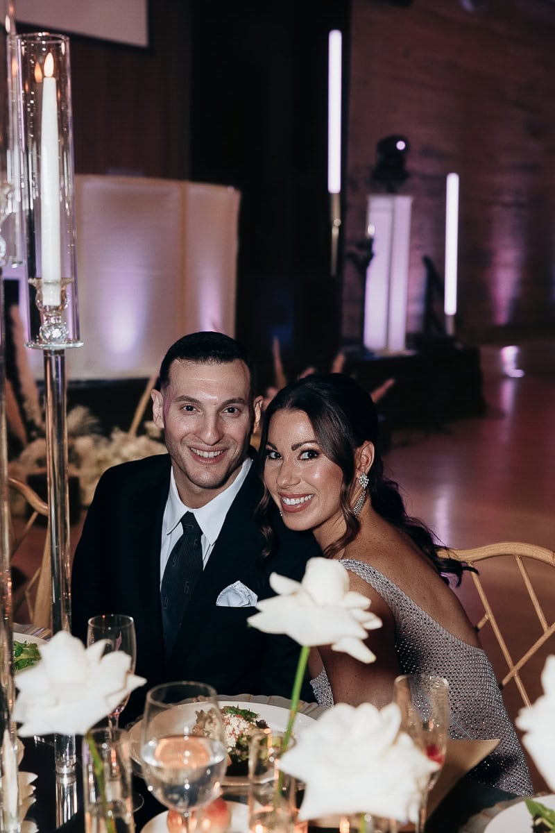 Couple seated at a candlelit reception table with white roses and purple uplighting at The Switch House by Cescaphe, Philadelphia, PA.