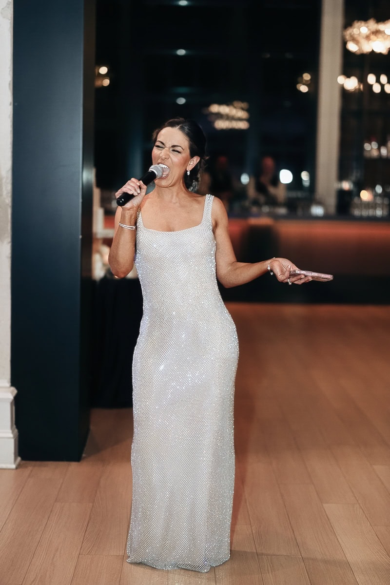 Woman in silver sequined gown delivering a mic toast on wood floor; chandeliers blur behind at The Switch House by Cescaphe, Philadelphia.