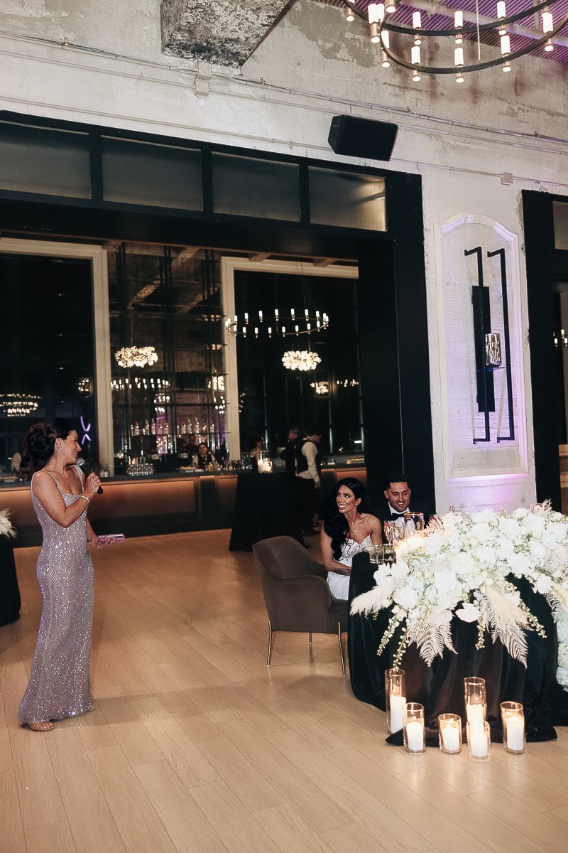 Guest in a silver sequin gown gives a toast as a couple sits at a candlelit sweetheart table with white florals in an industrial-chic hall at The Switch House by Cescaphe, Philadelphia.