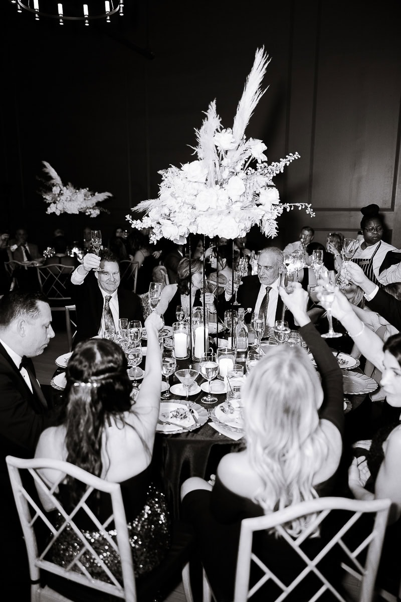 Black-and-white photo of guests raising champagne flutes around a round table with tall pampas floral centerpiece and candlelight at The Switch House.