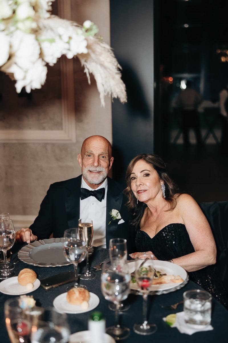 Two formal wedding guests seated at a dinner table with crystal glasses, silver charger, and bread rolls at The Switch House, Philadelphia.
