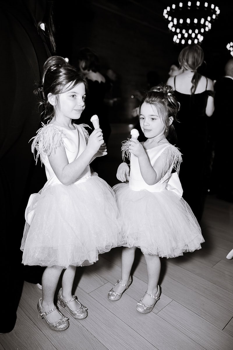 Two young girls in matching tulle dresses holding small ice cream cones at a wedding reception beneath blurred chandeliers, black-and-white.