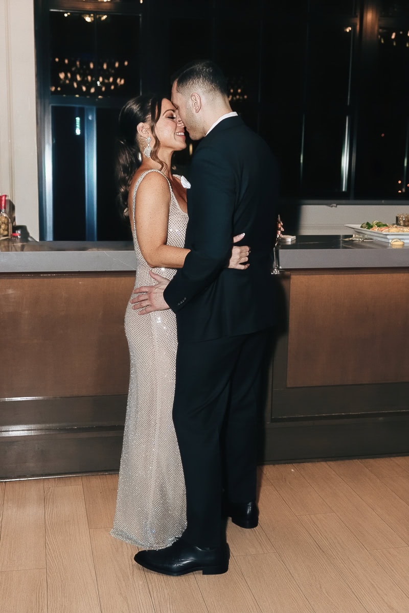 Couple in sequined gown and black suit embracing by the bar during evening reception; low light, champagne flute on counter.