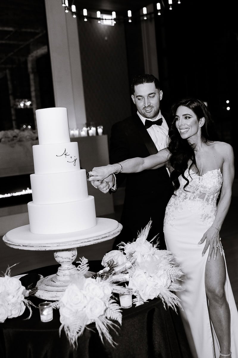 Couple cuts a four‑tier monogrammed cake on a pedestal amid roses and candles at The Switch House by Cescaphe, Philadelphia, PA.