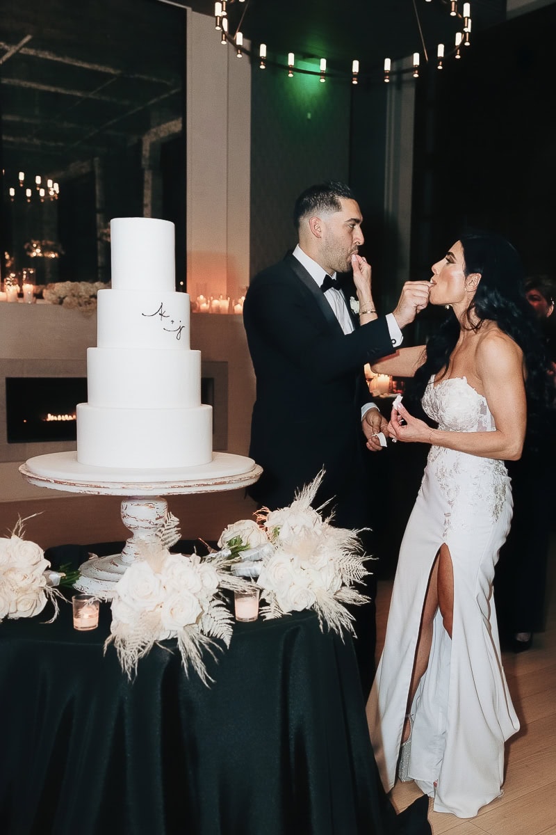 Couple feed each other cake beside four-tier white cake on black-draped table with white florals and candles at The Switch House, Philadelphia.