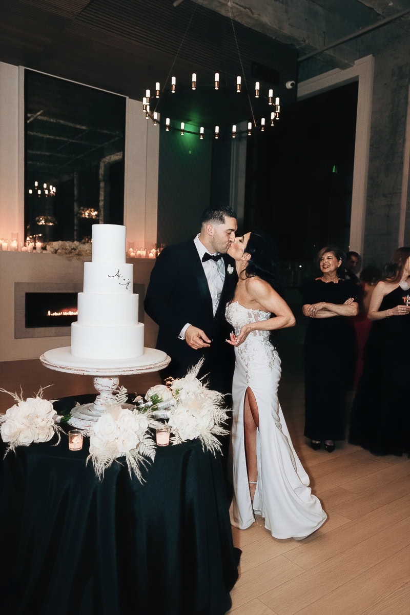 Couple kiss beside four-tier white cake on black-draped table with white florals and candles at The Switch House by Cescaphe, Philadelphia.