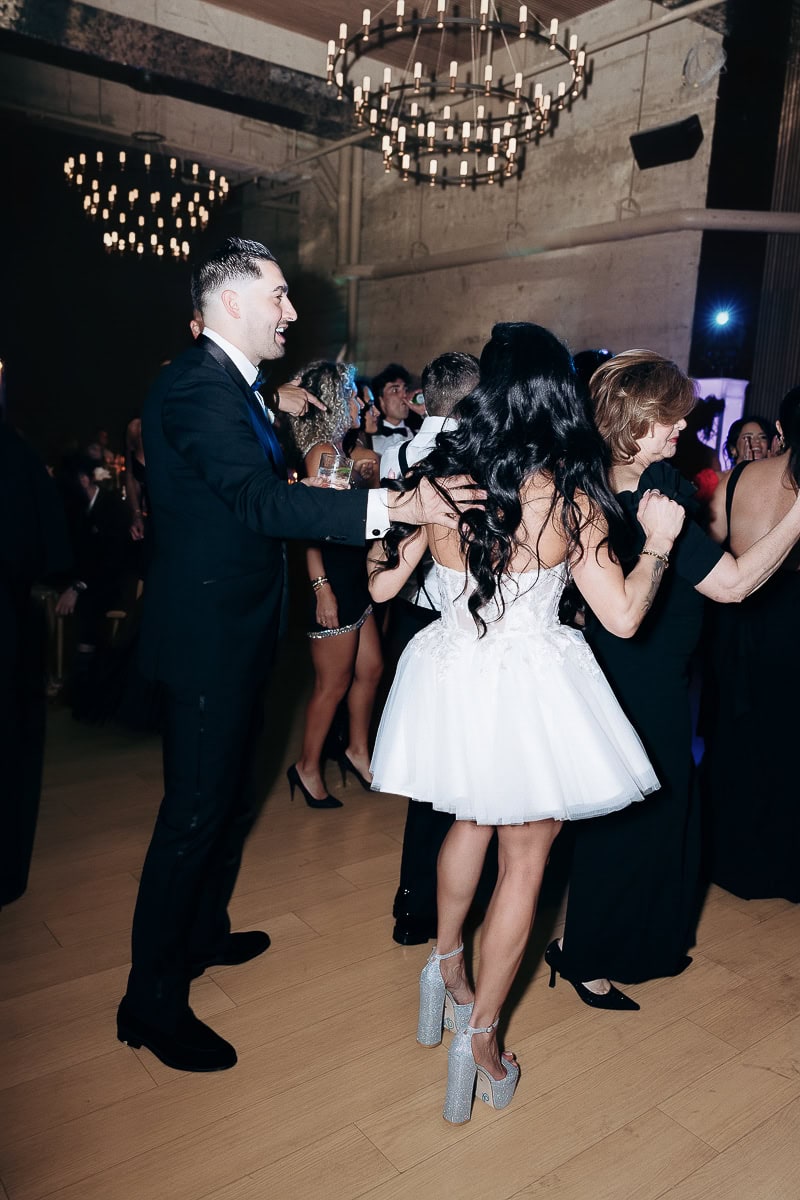 Black‑tie guests dance in a line; woman in white tulle mini and glitter platforms beneath chandeliers at The Switch House by Cescaphe, Philadelphia.