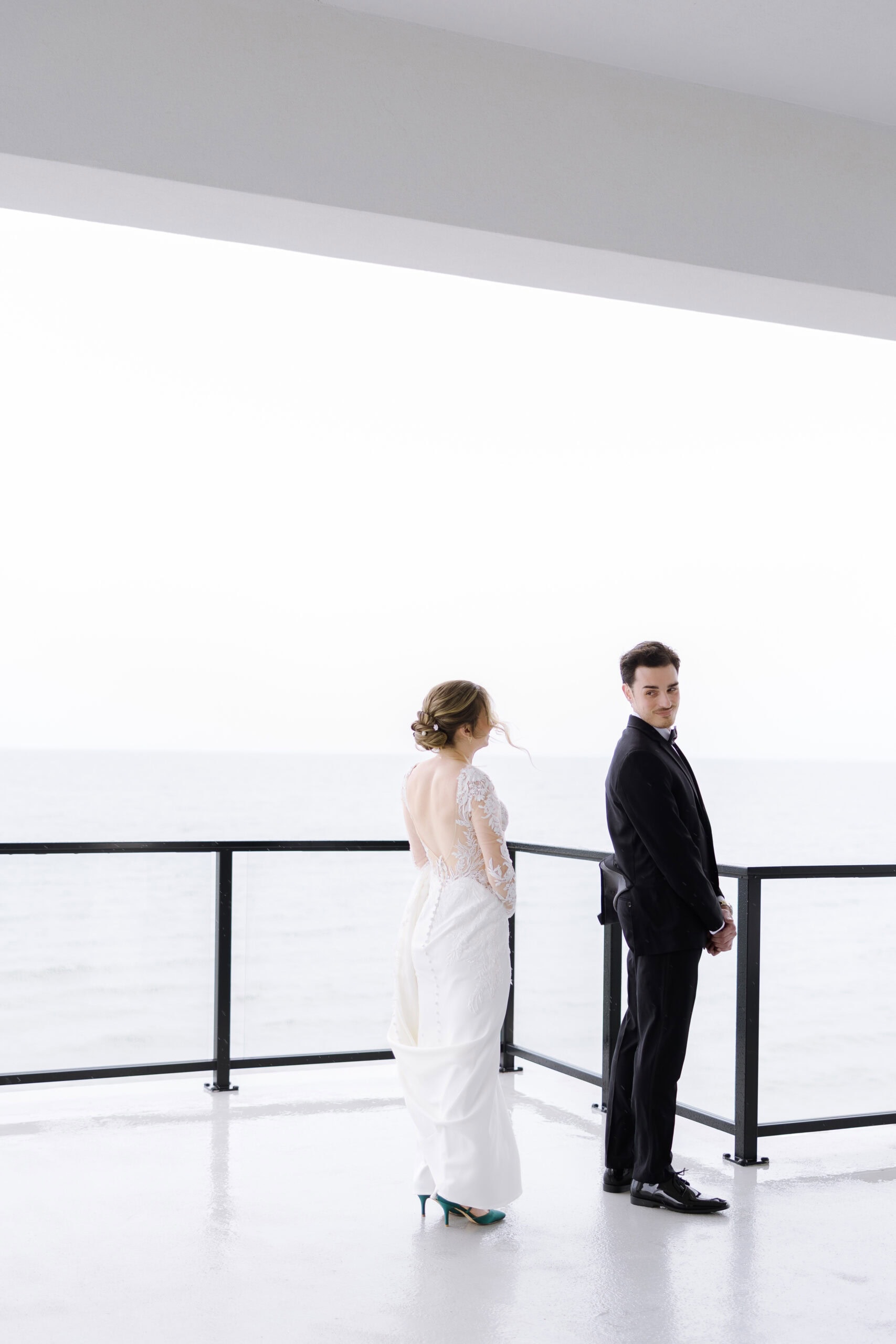 Bride in lace-back wedding dress and emerald heels with groom in black tuxedo on balcony overlooking ocean at Tyde at Walnut Beach, Milford, CT.