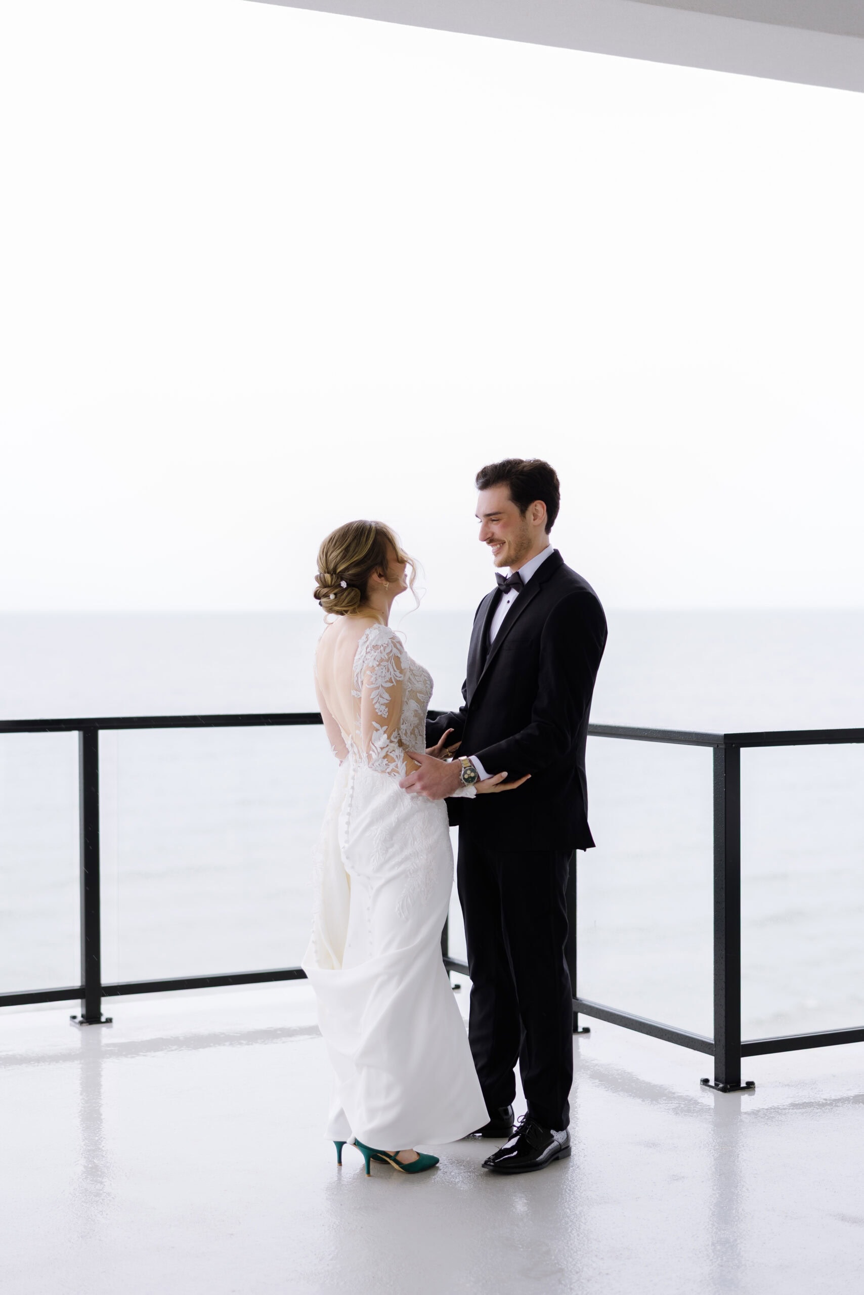 Bride in lace wedding dress and teal heels holding hands with groom in black tuxedo on balcony overlooking calm water at Tyde at Walnut Beach, Milford, CT.