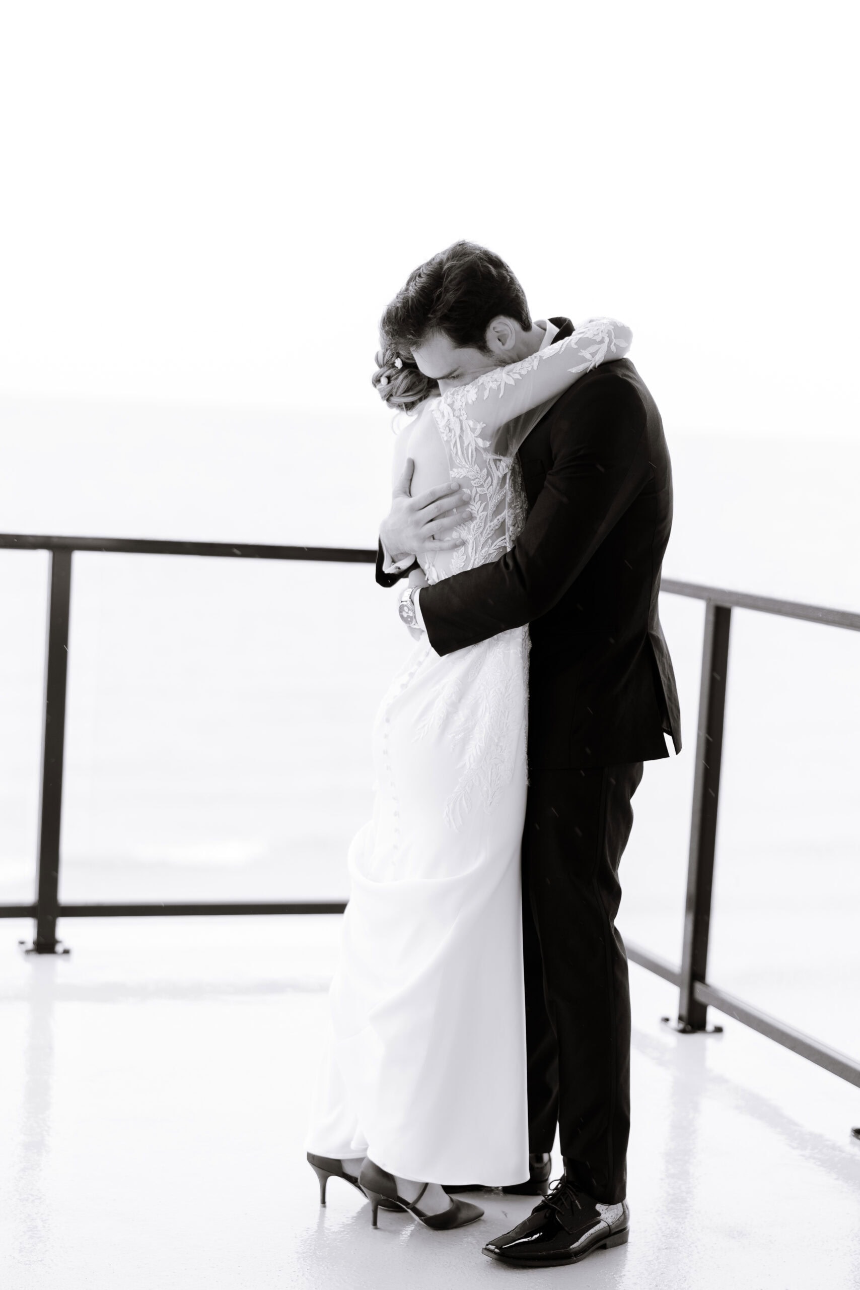 Black and white photo of a couple embracing on a balcony overlooking water, bride in lace gown and groom in black suit.