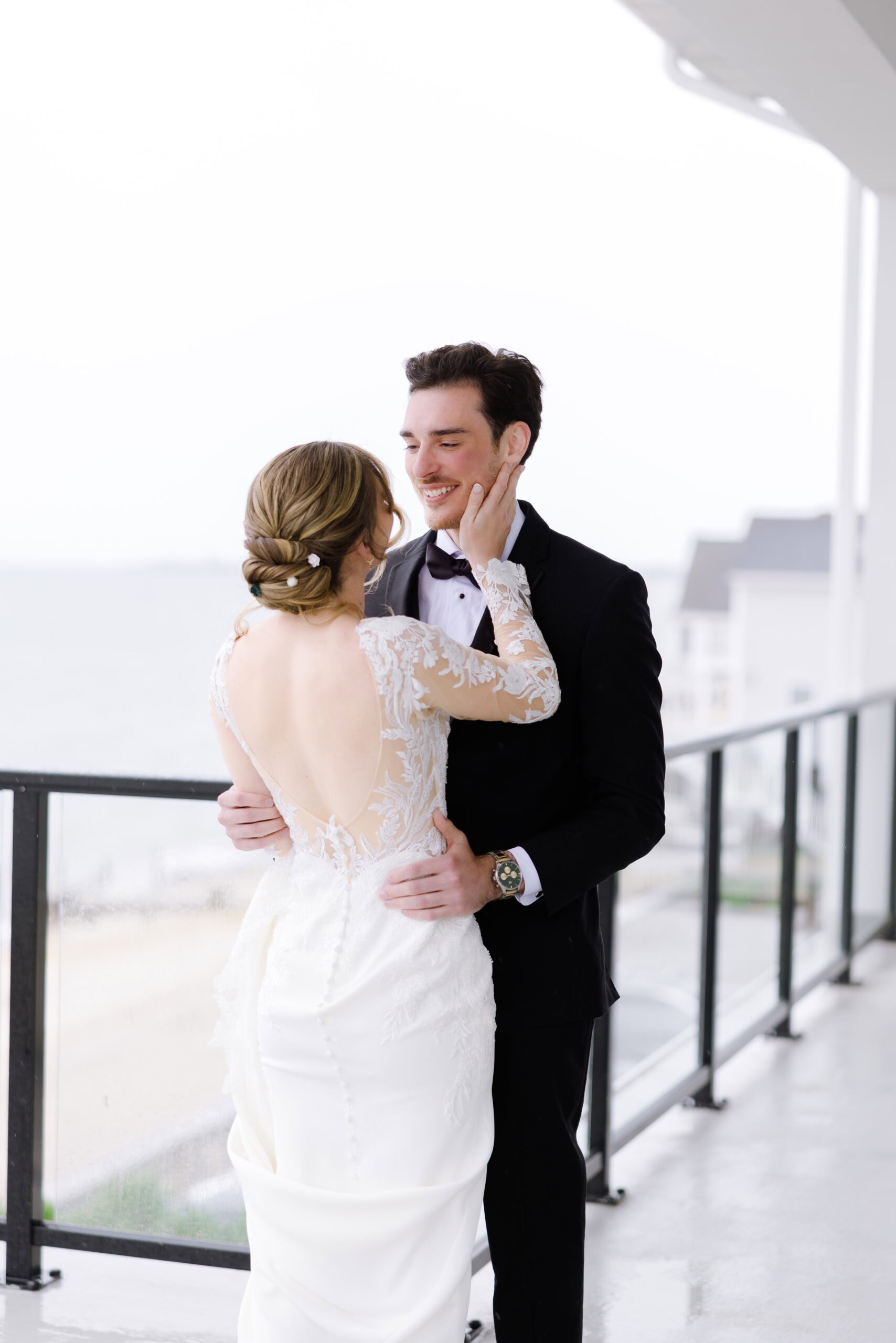 Bride in lace wedding dress with open back caressing groom's face on balcony overlooking water at Tyde at Walnut Beach, Milford, CT.