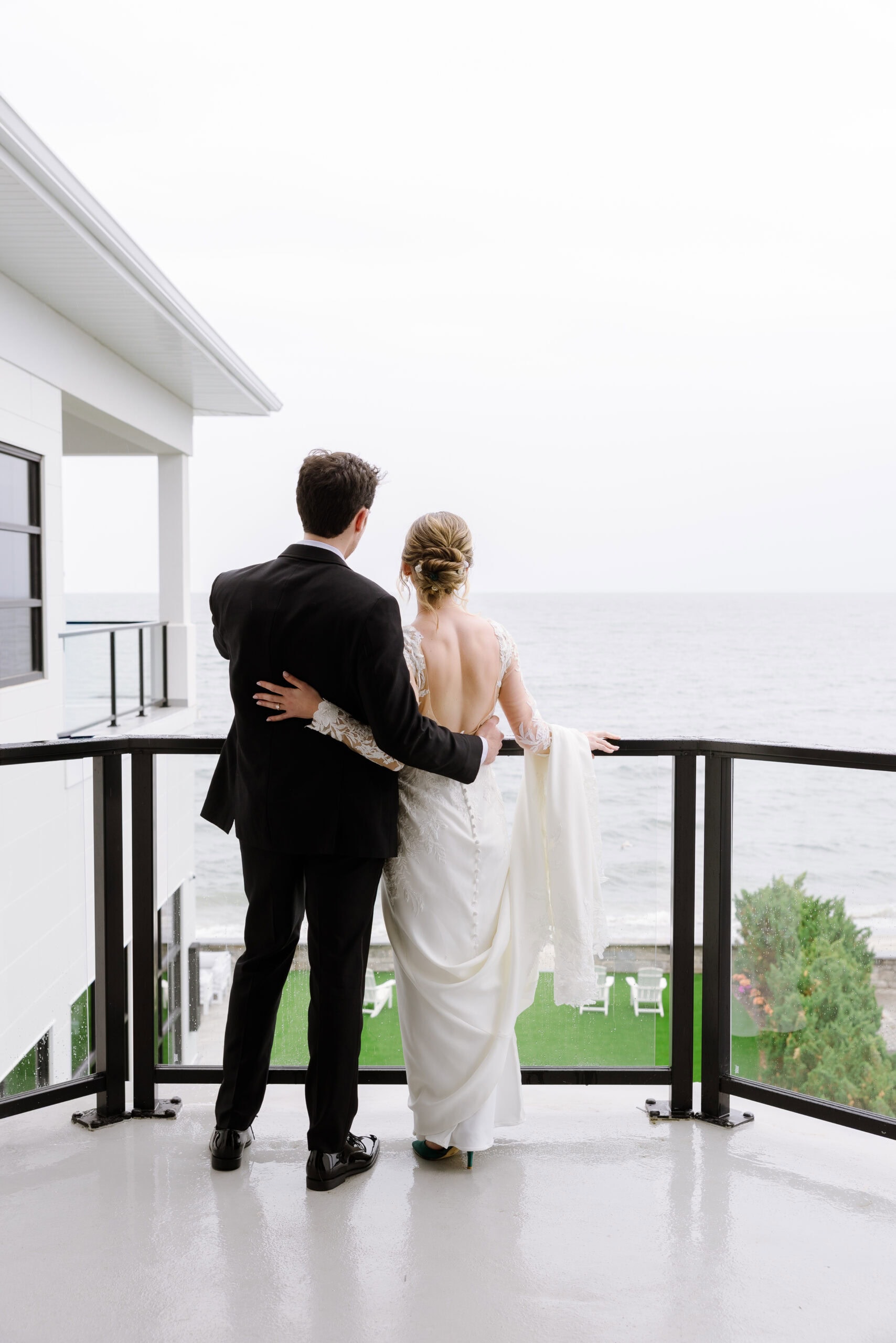 Bride and groom standing on balcony overlooking ocean, embracing with backs to camera at Tyde at Walnut Beach, Milford, CT.