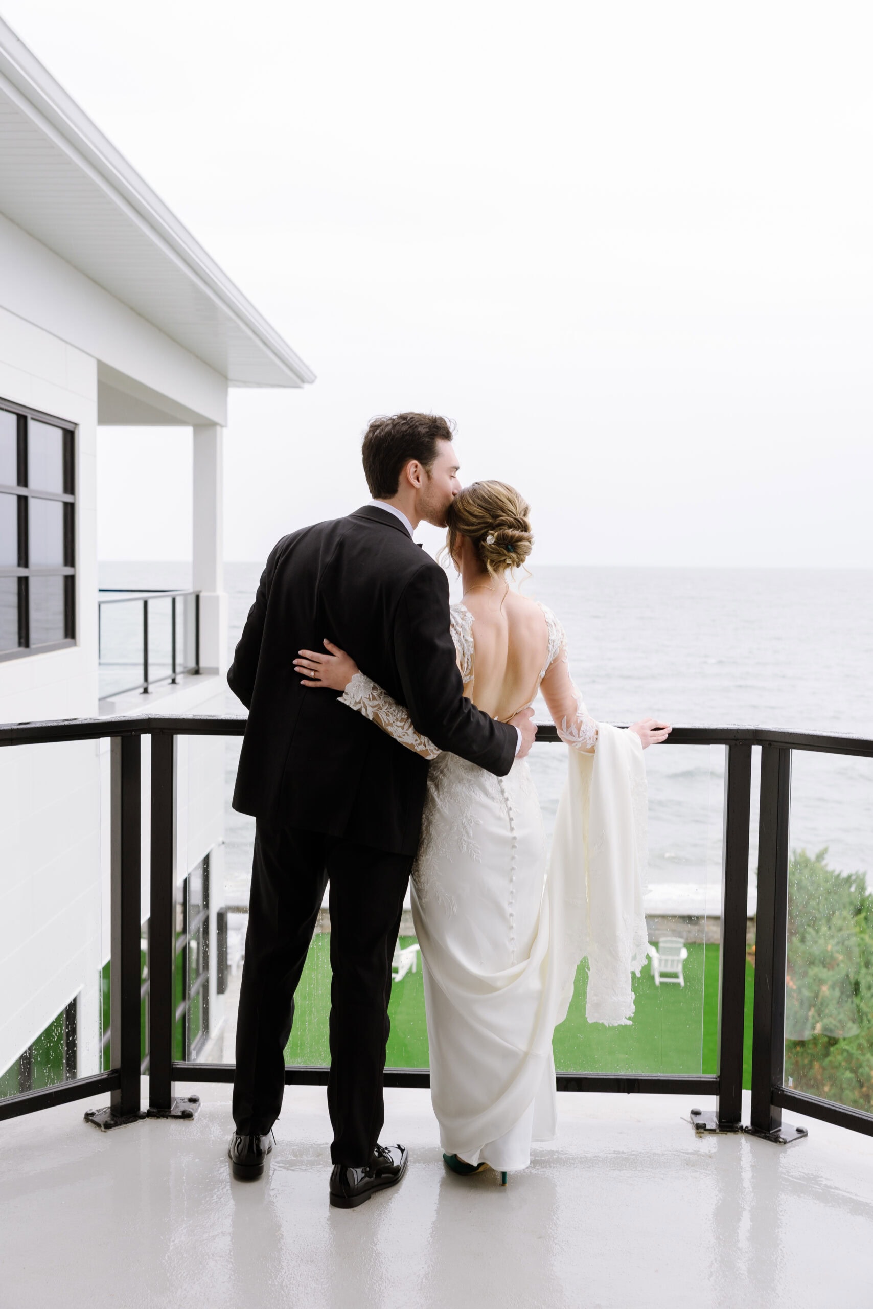 Bride and groom embracing on balcony overlooking ocean, groom kissing bride's temple, Tyde at Walnut Beach venue