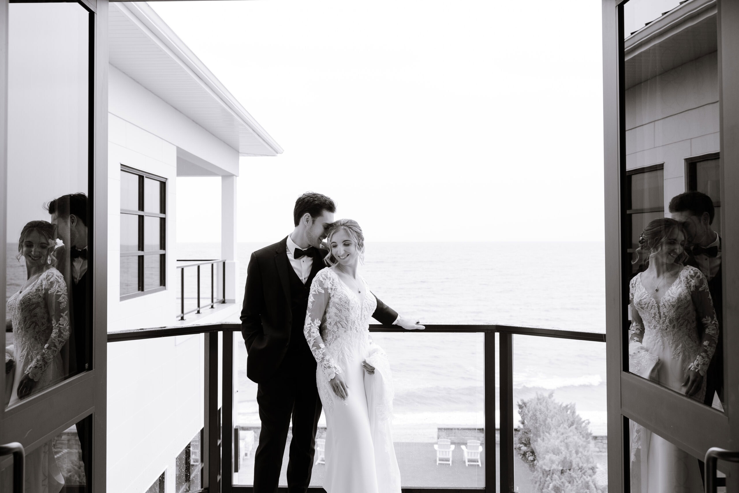 Black and white photo of a groom in tuxedo and bride in lace wedding dress embracing on a balcony overlooking the ocean, with reflections in glass doors.