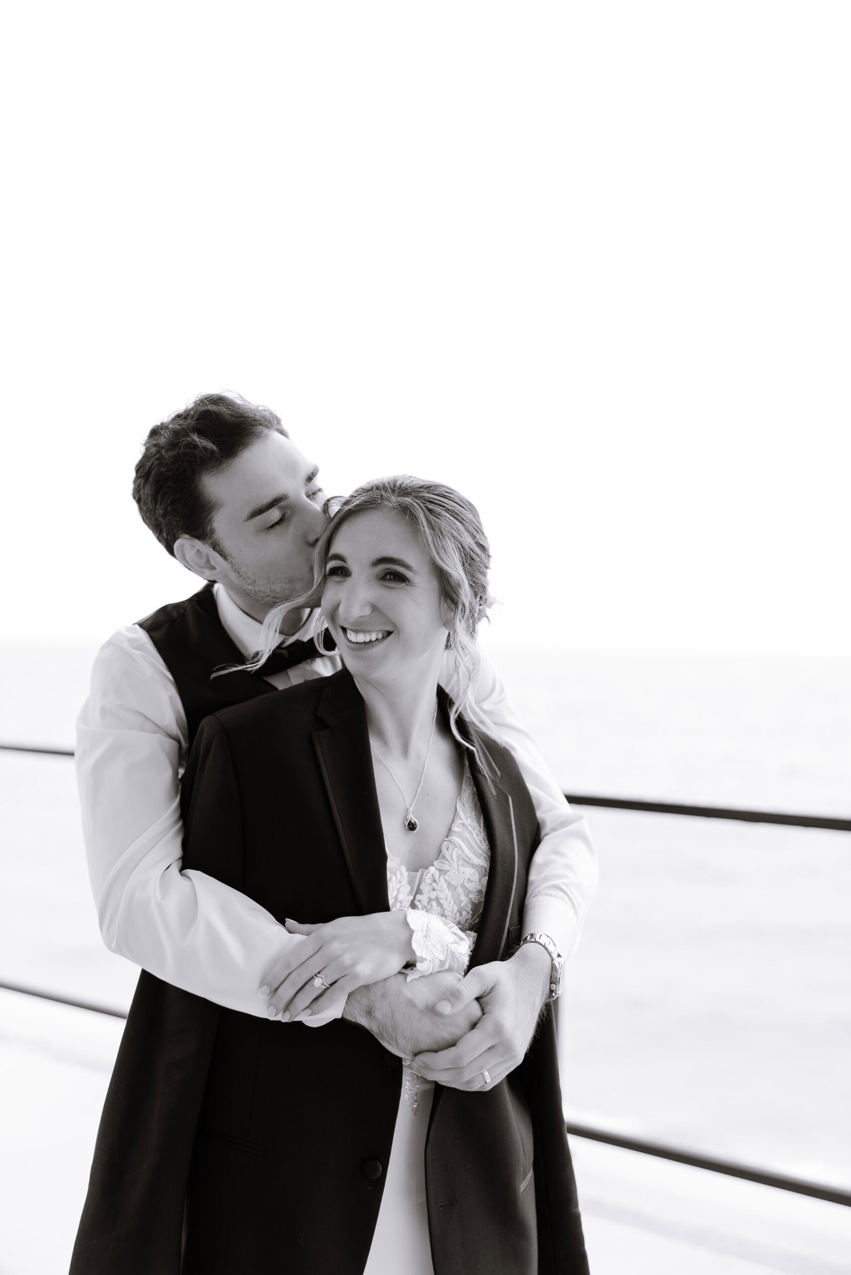 Black and white photo of a couple embracing by the ocean, the man kissing the woman's temple, both dressed in elegant wedding attire at Tyde at Walnut Beach, Milford, CT.