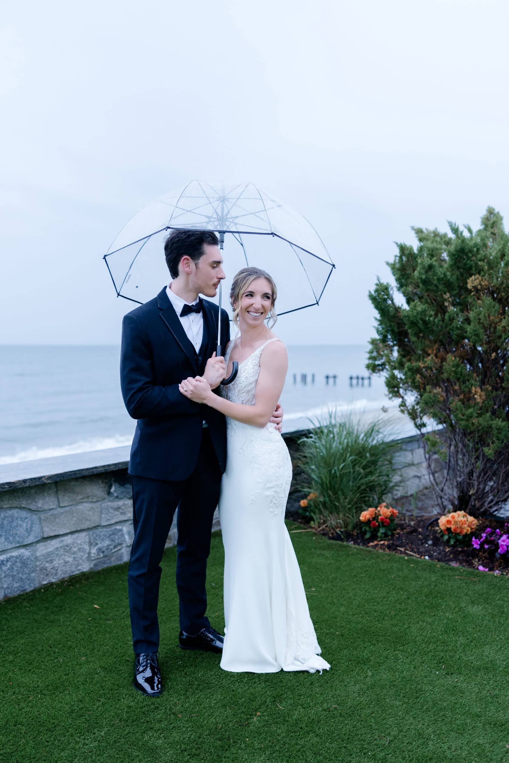Bride and groom standing close under a clear umbrella by the water's edge, with greenery and flowers nearby at Tyde at Walnut Beach, Milford, CT.