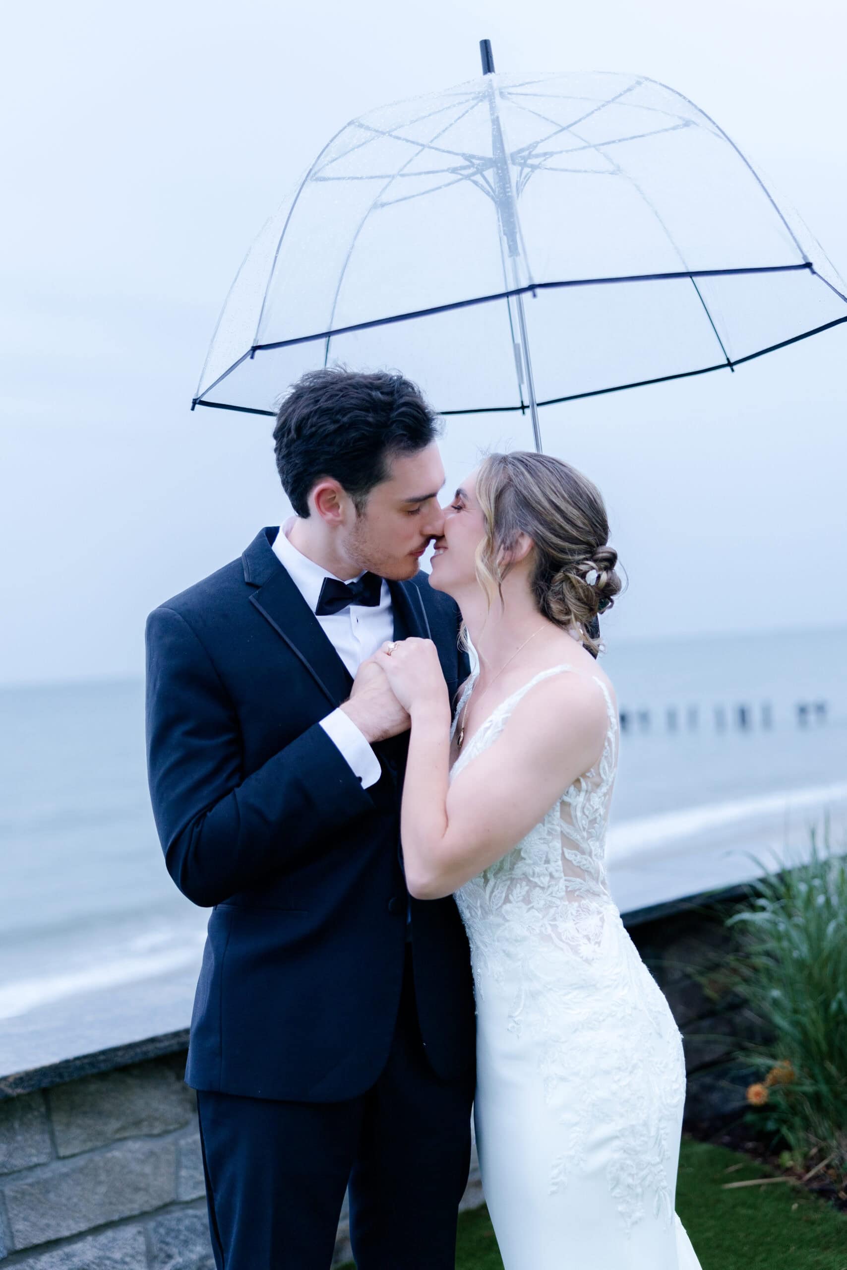 Couple kissing under a clear umbrella by the water at Tyde at Walnut Beach, Milford, CT, with bride in lace gown and groom in tuxedo.