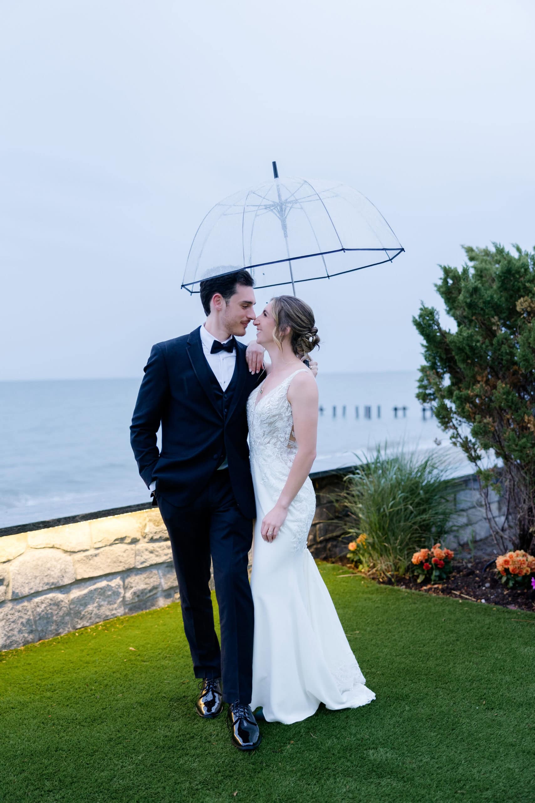 Bride and groom standing close under a transparent umbrella by the water at Tyde at Walnut Beach, Milford, CT, on a cloudy day.