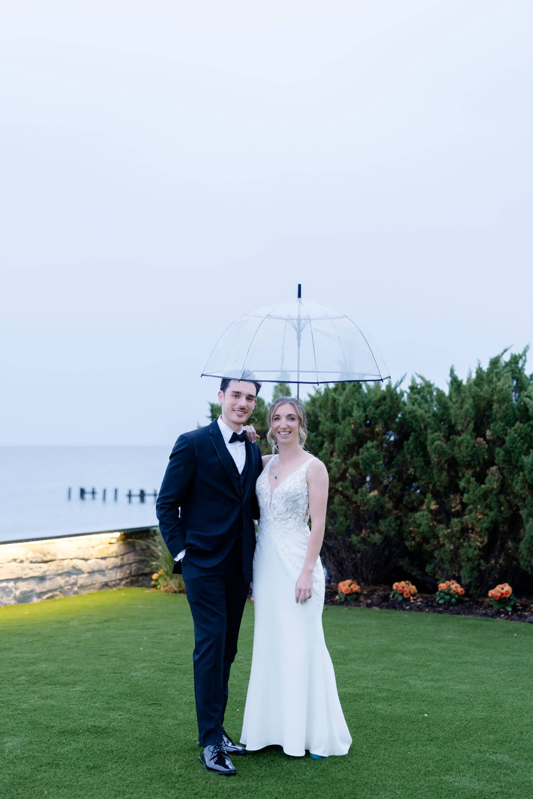 Wedding couple in formal attire standing on green lawn by the sea, sharing a transparent umbrella on an overcast day at Tyde at Walnut Beach, Milford, CT.