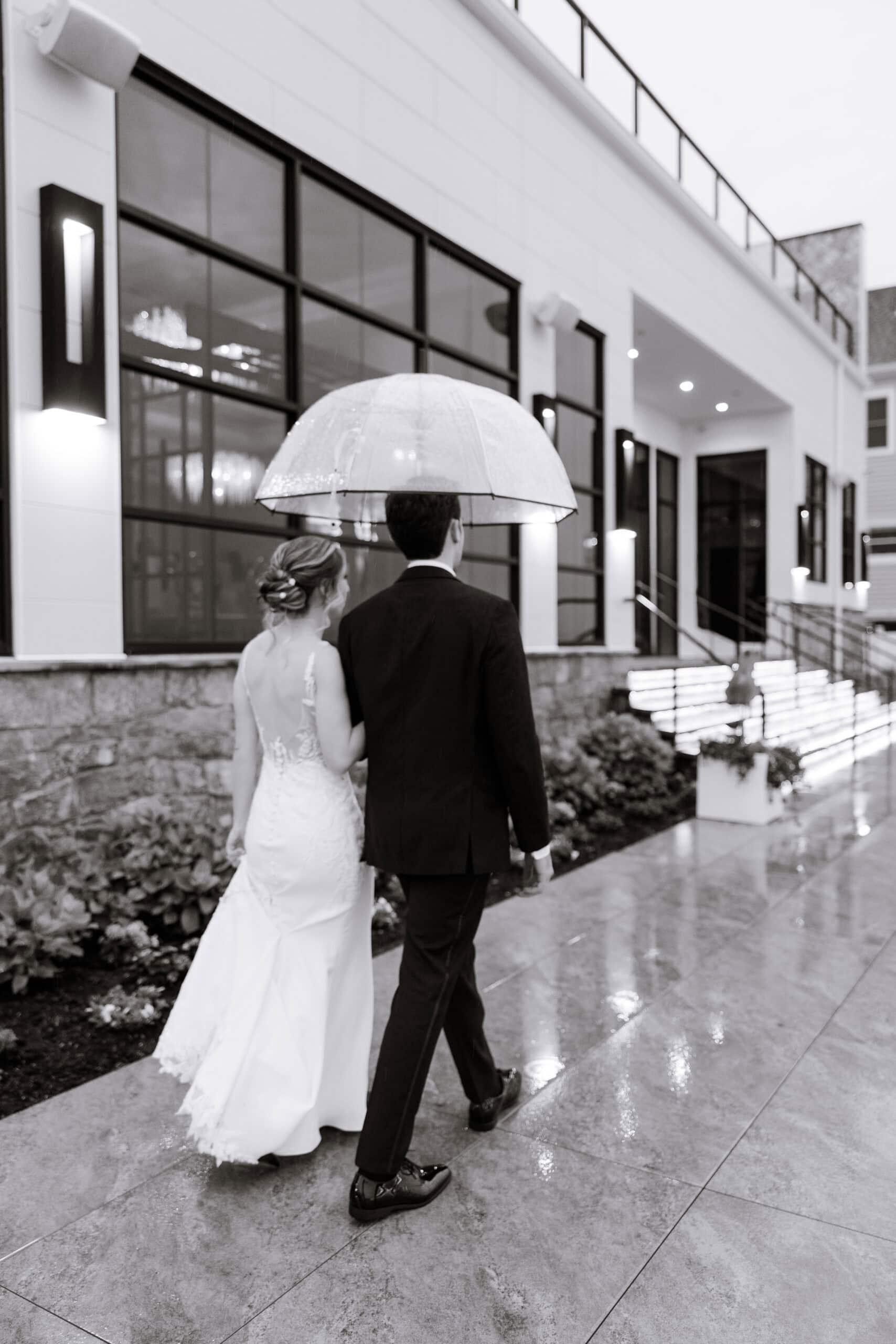 Bride and groom walking under a clear umbrella on a wet stone path outside a modern building at Tyde at Walnut Beach, Milford, CT.