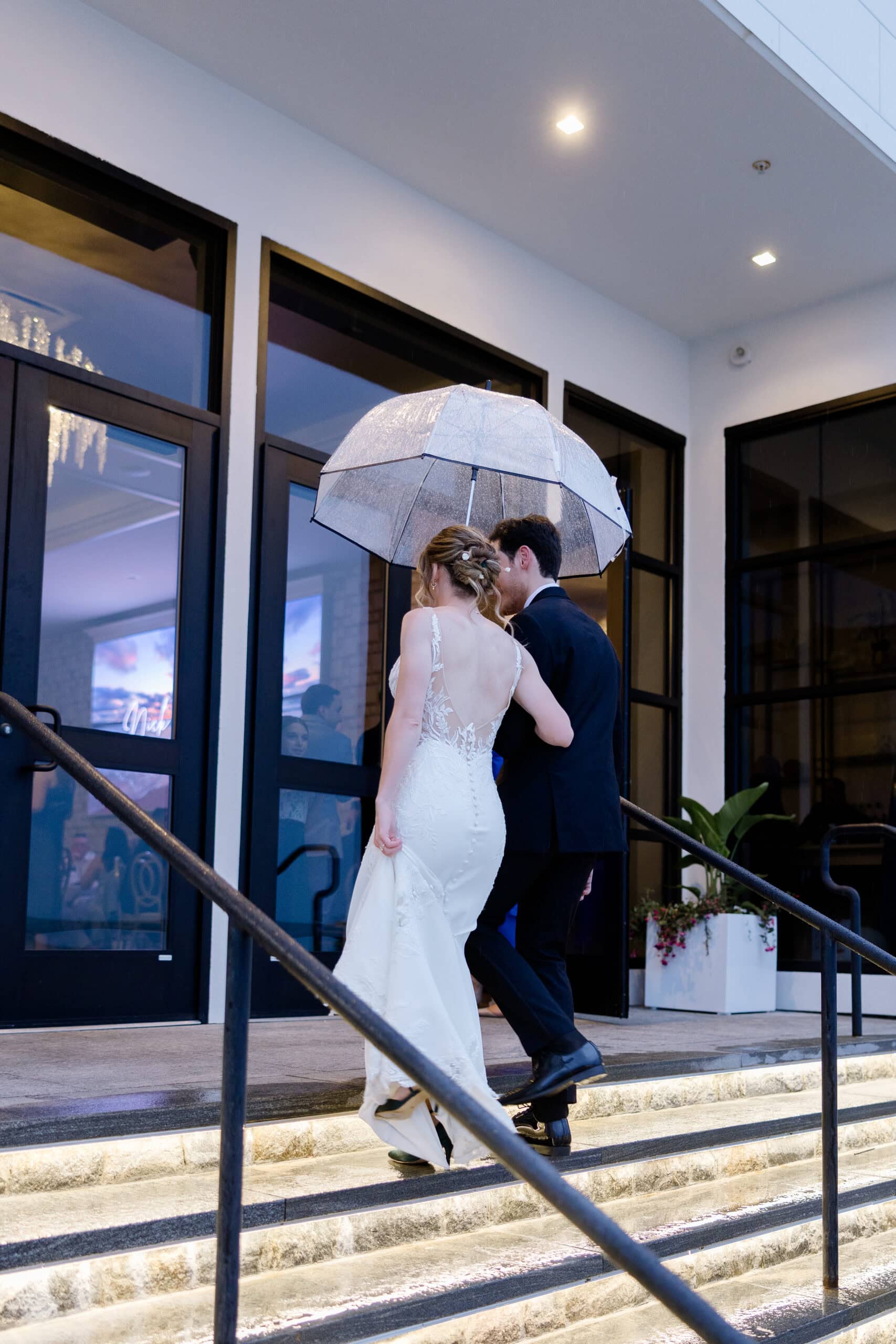 Bride and groom walking up outdoor steps under a transparent umbrella, evening light reflecting on wet stone, modern building entrance.