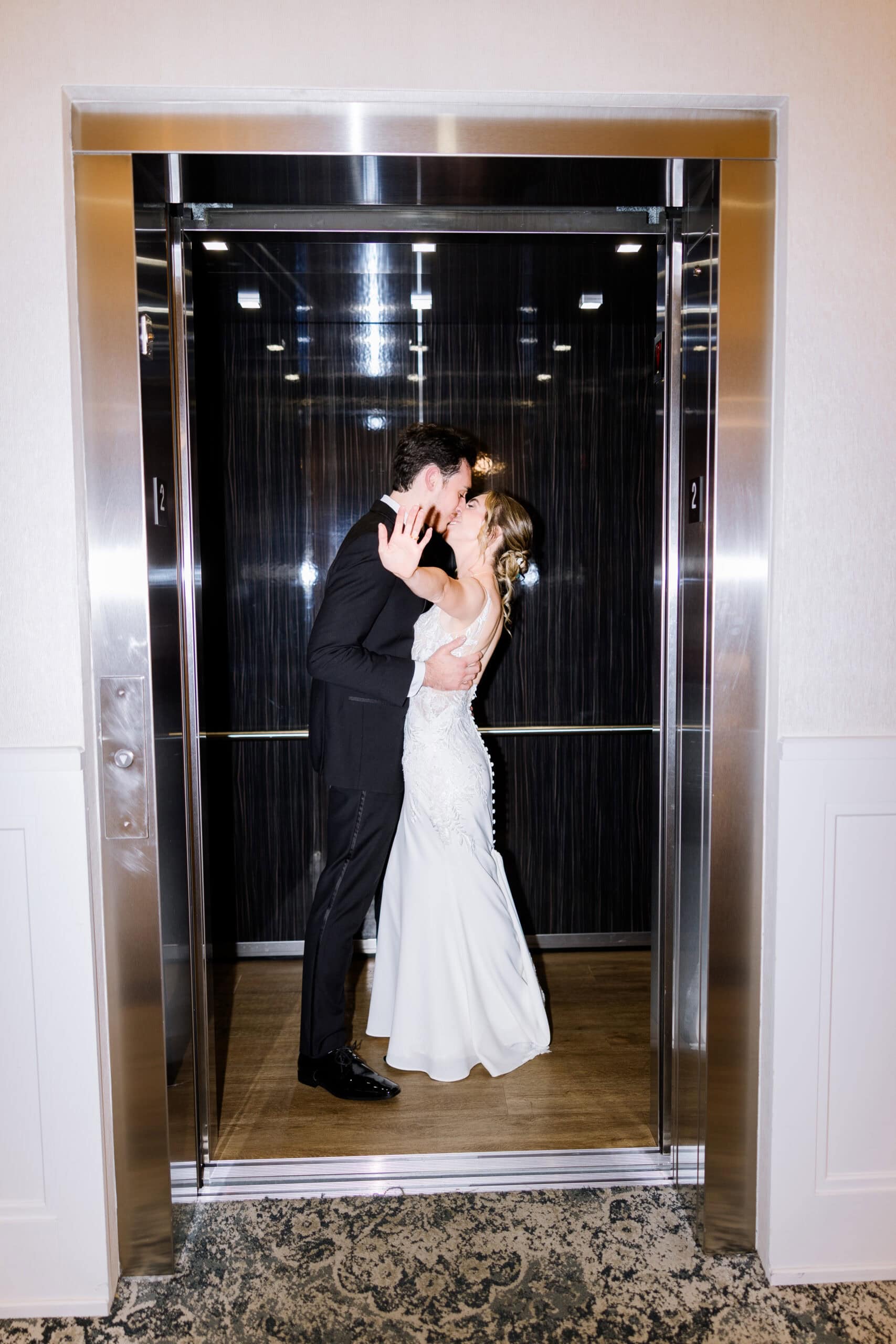 Bride and groom kissing inside a modern elevator with the bride's hand extended outward, showcasing a tender wedding moment.