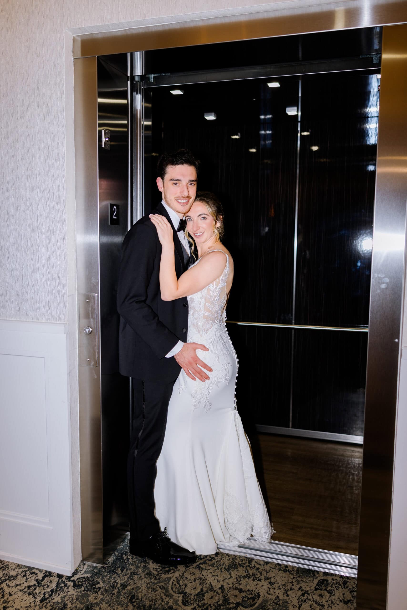 Bride in lace wedding gown and groom in black tuxedo embracing inside a modern elevator with reflective surfaces.